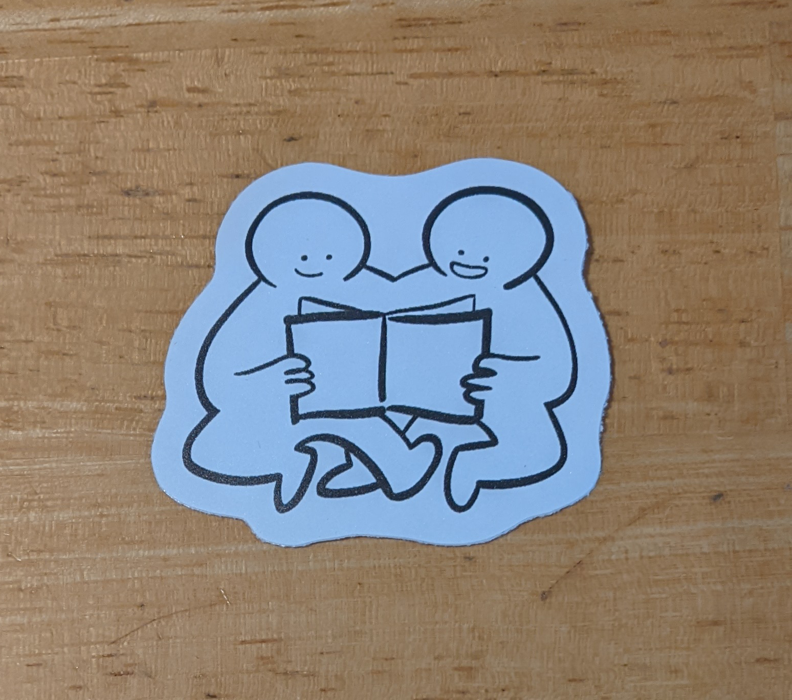 Black and white 5x5cm vinyl sticker of two friends sitting next to each other, smiling. They hold a book between them and read together.